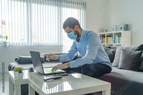 Young adult one man caucasian wearing gloves on hands and protective mask on face while working from home sitting by the table laptop preventing virus spread in epidemic quarantine health prevention