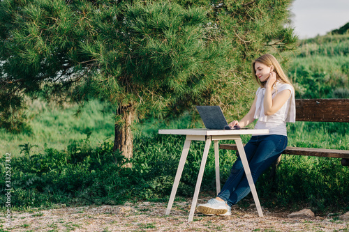 Young attractive freelance girl sitting on a bench and working on a laptop at a table outside near the tree
