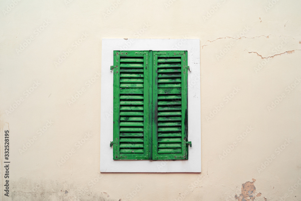 Old closed green wooden shutters on light plastered wall with cracks and time spots, in Mediterranean style. Outdoors, cozy vintage, copy space