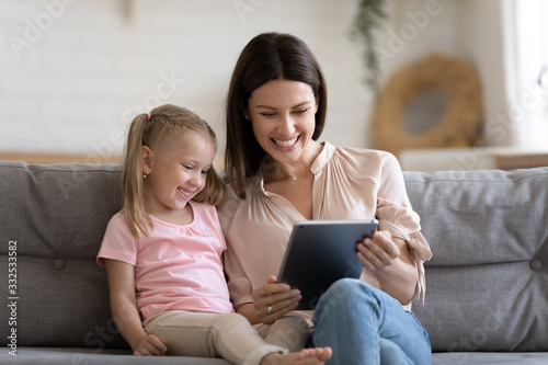 Smiling millennial nanny play with cute little girl child enjoy game on tablet relaxing on comfortable couch, happy mom and small daughter watch funny video cartoon on pad, spend weekend at home