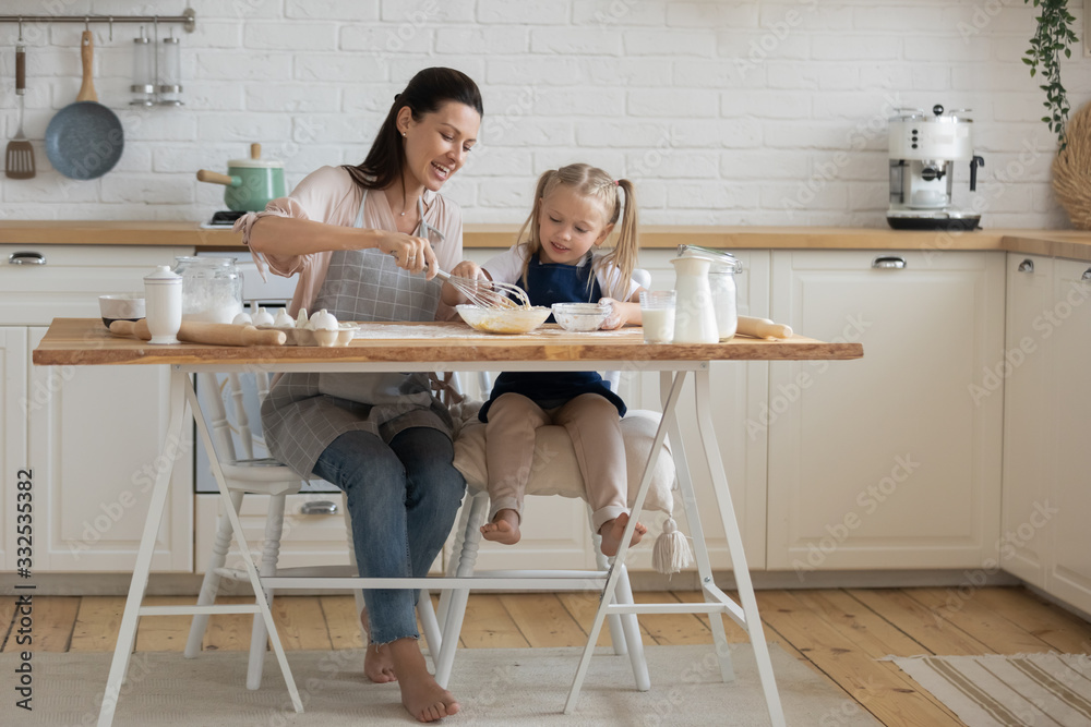 Young mother and little preschooler daughter sit at table in kitchen cooking breakfast together, smiling mom teach small girl daughter whisk eggs prepare pancakes or cookies, family recipe concept Young mother and little preschooler daughter sit at table in kitchen cooking breakfast together, smiling mom teach small girl daughter whisk eggs prepare pancakes or cookies, family recipe concept