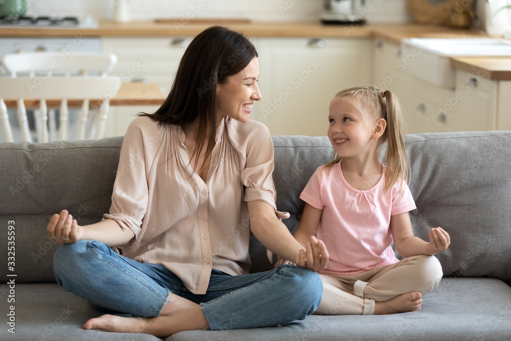 Smiling active young mom and preschooler daughter sit on couch practicing yoga in lotus position, happy funny mother and girl child meditate with mudra hands have fun, follow healthy lifestyle Smiling active young mom and preschooler daughter sit on couch practicing yoga in lotus position, happy funny mother and girl child meditate with mudra hands have fun, follow healthy lifestyle