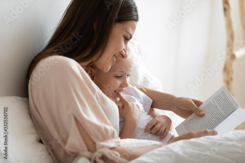 Close up of smiling mom and little daughter relax in cozy bed read funny interesting children book together, happy mother and small preschooler girl child enjoy fairytale rest in comfortable bedroom