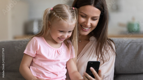 Close up of smiling young mother and small preschooler daughter relax at home together watch funny cartoon on smartphone, happy mom and little girl child have fun using cellphone playing