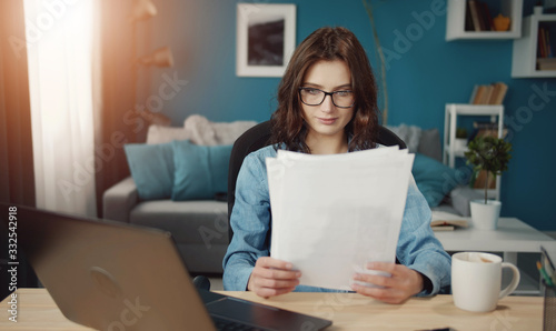 Focused young woman working at home looking at papers sitting at desk next to laptop computer