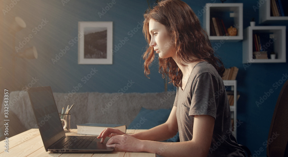 Young serious woman in casual wear using laptop sitting at desk in living room, side view Young serious woman in casual wear using laptop sitting at desk in living room, side view