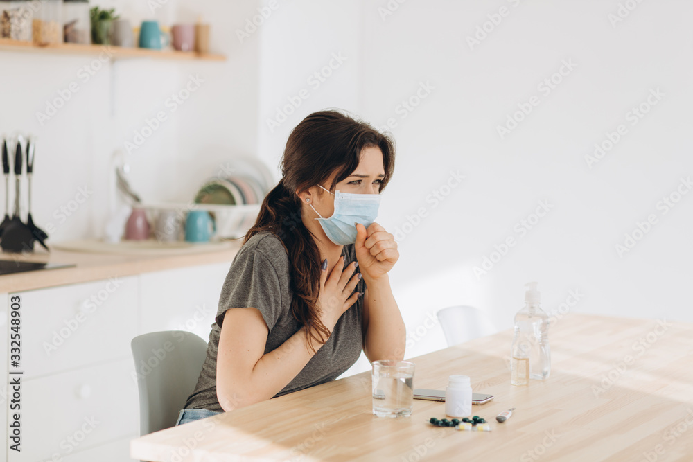 Woman in protective mask coughing in fist sick of coronavirus viral infection spreading coronavirus covering mouth and nose. Painful cough ill patient sitting in kitchen at home quarantine Woman in protective mask coughing in fist sick of coronavirus viral infection spreading coronavirus covering mouth and nose. Painful cough ill patient sitting in kitchen at home quarantine
