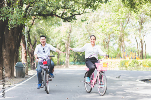 Thai Asian couples are riding bicycles in the park happily and joyfully on weekends, with clear skies, fresh air and green tree backdrop.