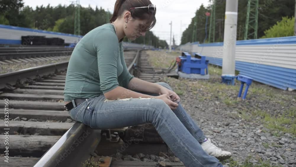 Young beautiful woman sits on the rails of the railway and uses a smartphone on a cloudy summer day Young beautiful woman sits on the rails of the railway and uses a smartphone on a cloudy summer day