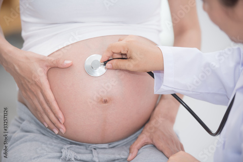 Young preganant woman expecting baby doctor visit. pregnancy, medicine, healthcare and people concept. gynecologist doctor with stethoscope listening to pregnant asian woman baby heartbeat at hospital photo