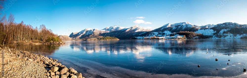 Panorama of nearly frozen Oltedalsvatnet lake and coastline with mountains peaks covered by snow during winter season, Gjesdal commune, Norway, February 2018