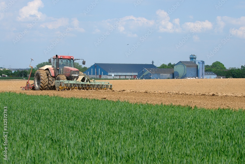Fototapeta premium Semis mais avec semoir 32 rangs de 4m et outil ottavioli (tasseligne) à l'avant. Coopérative agricole en arrière plan