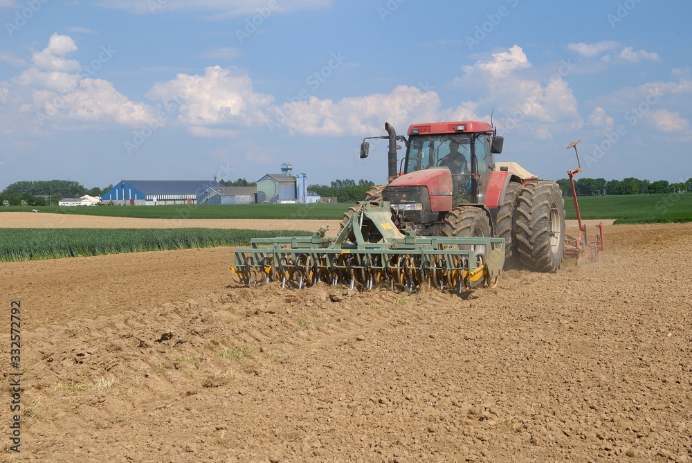 Fototapeta premium Semis mais avec semoir 32 rangs de 4m et outil ottavioli (tasseligne) à l'avant. Coopérative agricole en arrière plan