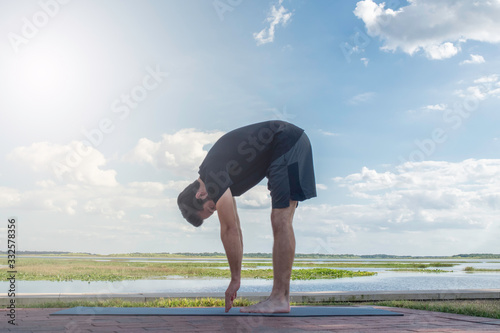Man wearing a black shirt and dark green shorts bends over in a forward fold position correcting his bodies posture in front of a bright blue sky with gorgeous white clouds behind his body.