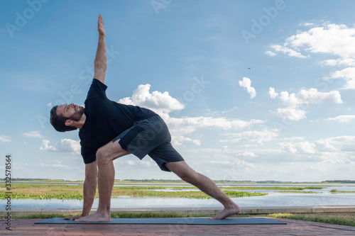 Man wearing a black shirt and dark green shorts bends over in a triangle pose correcting his bodies posture in front of a bright blue sky with gorgeous white clouds behind his body.