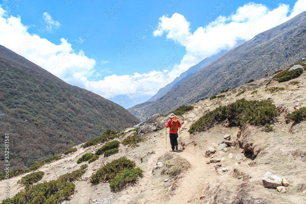 Back view. Man in red windbreaker with orange backpack walks on footpath in Himalayas on the way back from Everest base camp. Clouds lies on mountainside. Theme of trekking in Nepal.