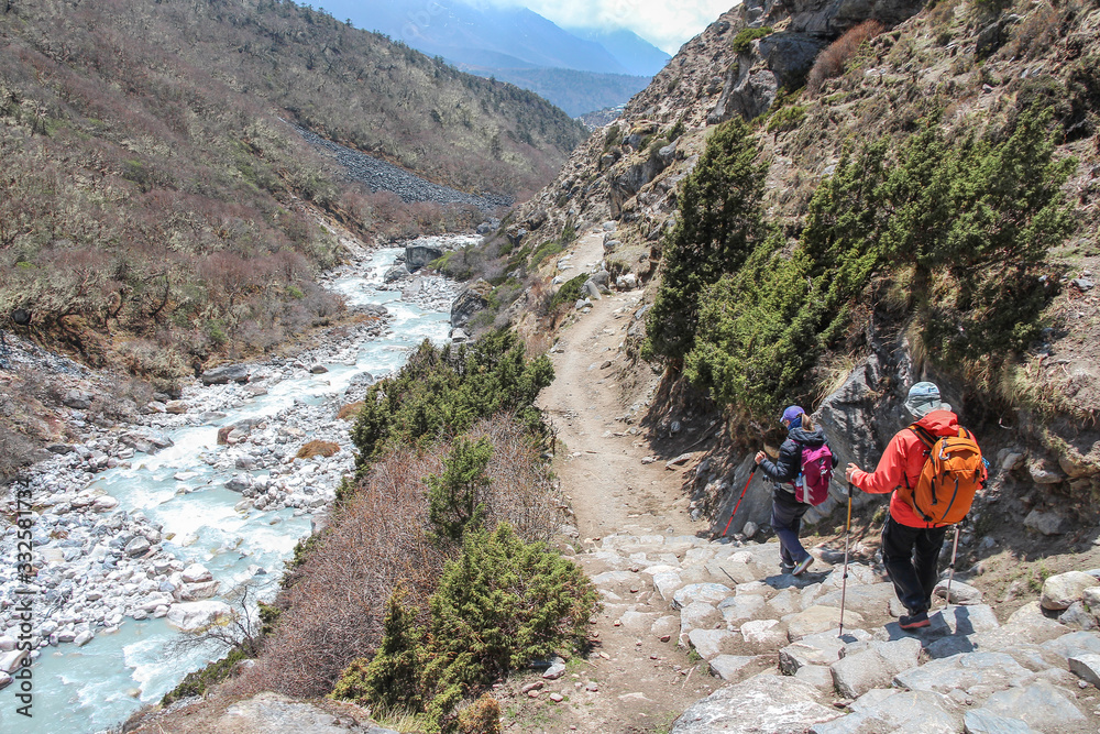Back view. Couple of hikers with backpacks walks on footpath in Himalayas on the way back from Everest base camp. Imja Khola river flows near footpath. Theme of trekking in Nepal.