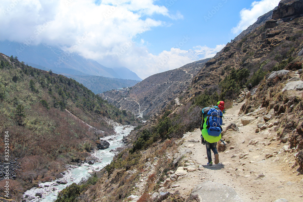 Back view. Some nepalese sherpa porters with backpacks walks on footpath in Himalayas on the way back from Everest base camp. Imja Khola river flows near footpath. Theme of trekking in Nepal.