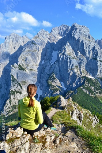 Young girl is sitting on the top of the mountains, enjoying beautiful view on the mountain range Kaisergebirge, Austria. Sunny day, white clouds, blue sky. 