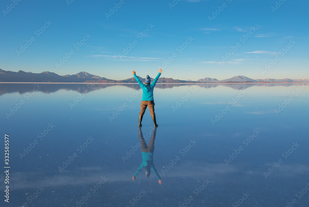Sunrise on Salar de Uyuni in Bolivia covered with water, woman tourist on salt flat desert and sky reflections
