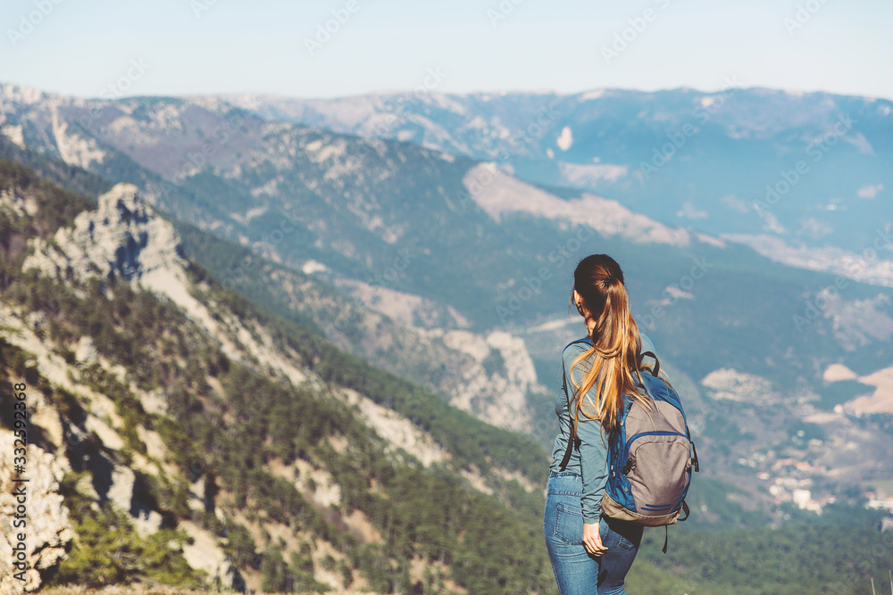 Young beautiful girl travels alone in the mountains in spring or autumn, looks into the distance and enjoys nature, rocks and green forests, view of the landscape. a backpack behind and sportswear