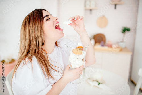 Happy plus size model in jeans and a white T-shirt, holding a sweetness in her hands, opening her mouth in a stylish bright kitchen. The body is positive. The concept of one beauty. XXXL fashion