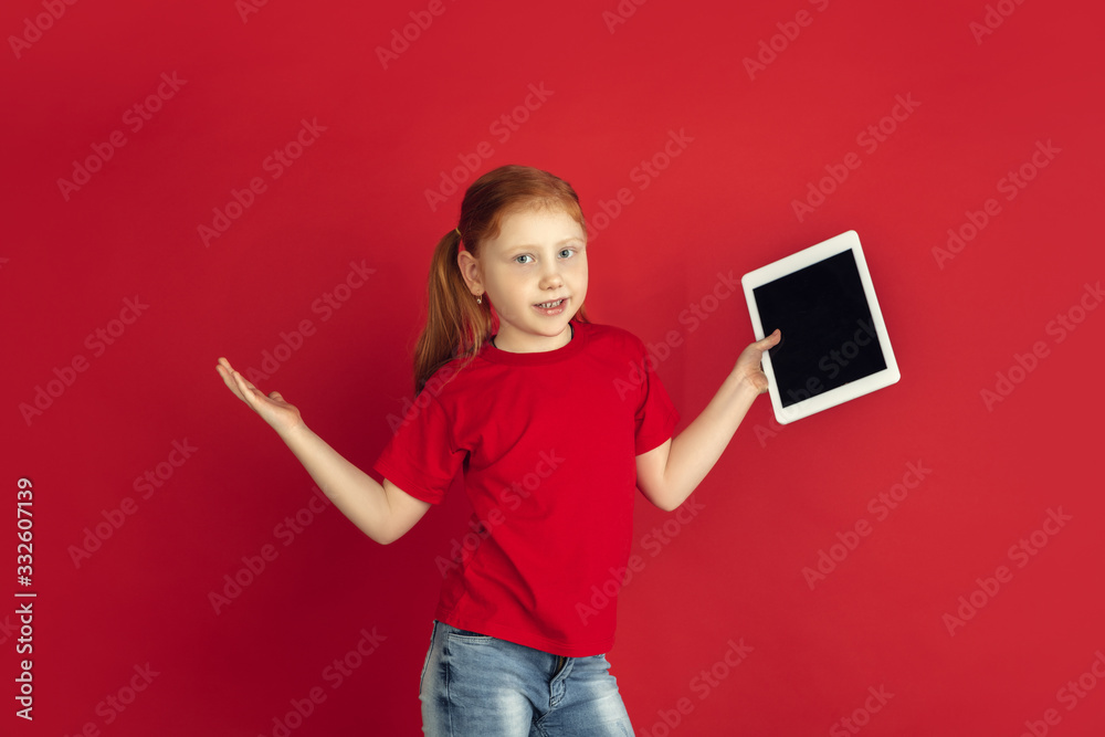 Using tablet, wondered. Caucasian little girl portrait isolated on red studio background. Cute redhair model in red shirt. Concept of human emotions, facial expression, sales, ad, childhood. Copyspace Using tablet, wondered. Caucasian little girl portrait isolated on red studio background. Cute redhair model in red shirt. Concept of human emotions, facial expression, sales, ad, childhood. Copyspace