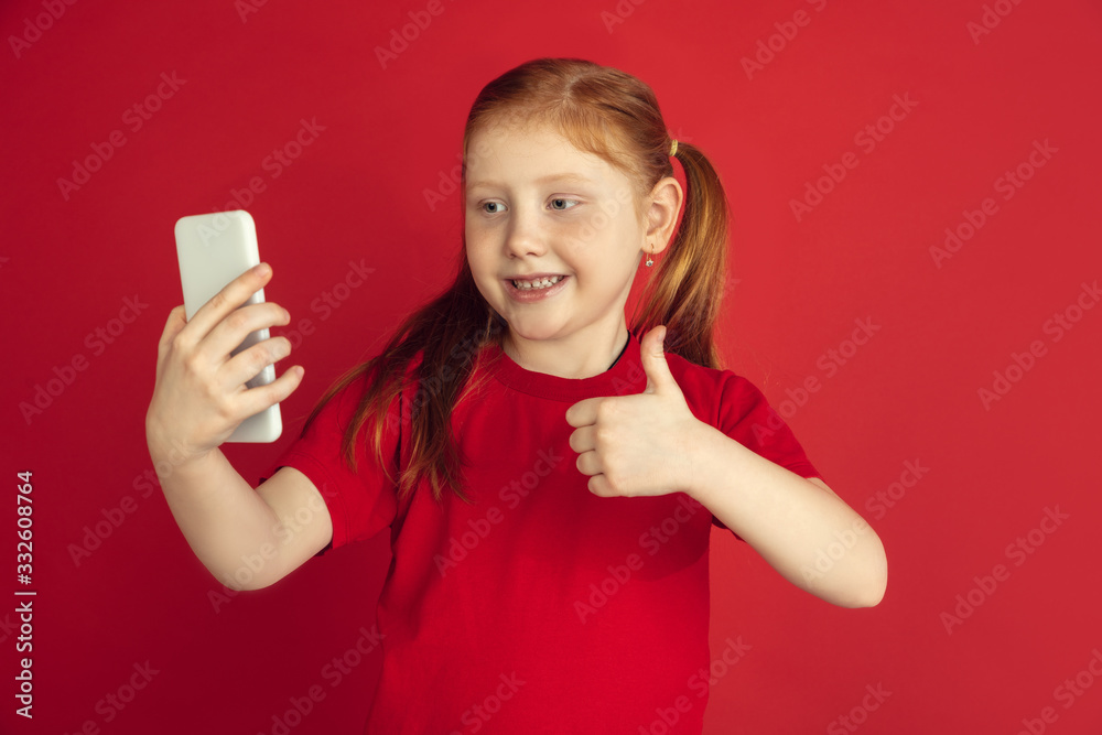Taking selfie, vlog. Caucasian little girl portrait isolated on red studio background. Cute redhair model in red shirt. Concept of human emotions, facial expression, sales, ad, childhood. Copyspace. Taking selfie, vlog. Caucasian little girl portrait isolated on red studio background. Cute redhair model in red shirt. Concept of human emotions, facial expression, sales, ad, childhood. Copyspace.