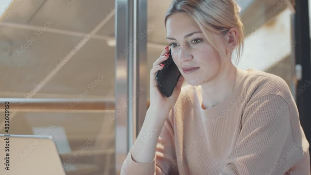 Tilt down shot of young female manager smiling and speaking on phone while working on laptop at desk in office with glass walls