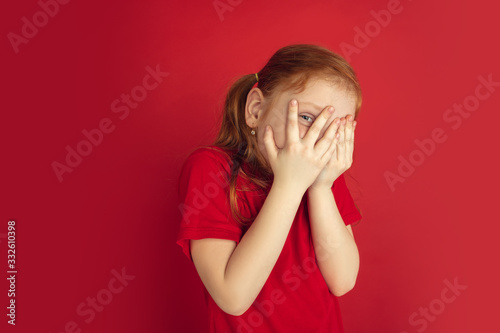 Scared hiding face. Caucasian little girl portrait isolated on red studio background. Cute redhair model in red shirt. Concept of human emotions, facial expression, sales, ad, childhood. Copyspace. © master1305