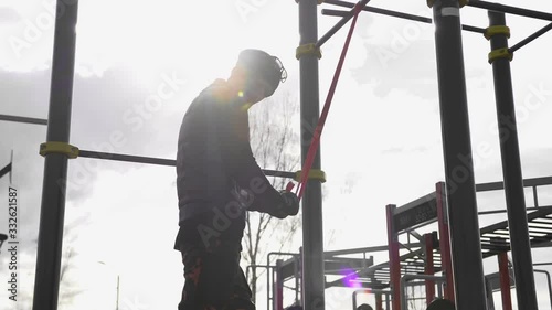 Man, black clothes hat glasses early, morning in spring, in sunny weather glare from sun on outdoor sports ground. Elastic elastic fixed horizontal bar athlete makes approaches shakes triceps muscles.
