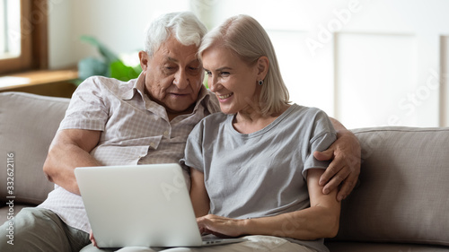 Smiling older wife and husband hugging, using laptop, sitting on cozy couch, watching movie or shopping online, looking at screen, mature man and woman spending leisure time together at home