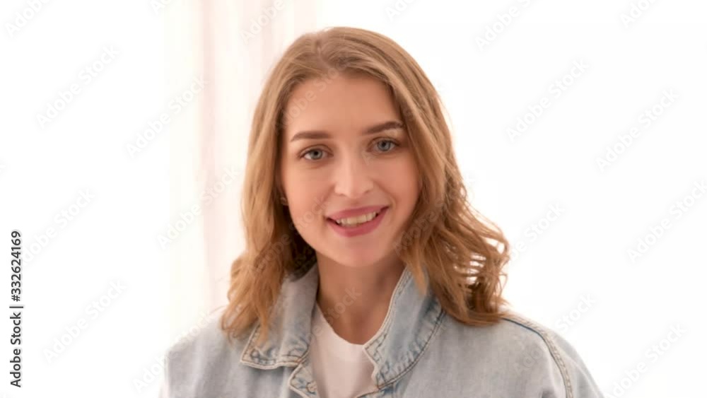Millennial female in casual clothes white shirt and denim jacket smiling at camera looking at camera and speaking against white wall in light room Millennial female in casual clothes white shirt and denim jacket smiling at camera looking at camera and speaking against white wall in light room