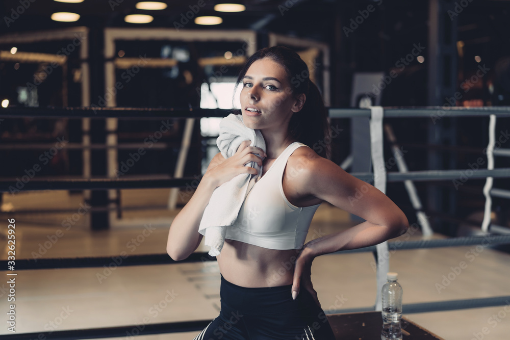 Smiling fit girl holding towel and taking rest in gym. Girl wipes sweat with a towel