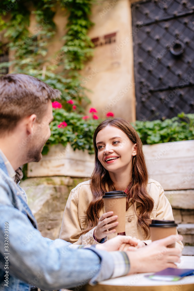 Obraz premium Selective focus of couple with paper cups of coffee looking at each other in cafe in city