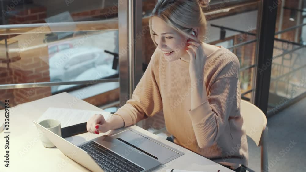 High angle shot of attractive blonde woman in wireless earphones smiling and chatting via video call on laptop while working at desk in loft office with glass walls High angle shot of attractive blonde woman in wireless earphones smiling and chatting via video call on laptop while working at desk in loft office with glass walls