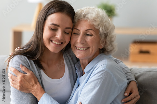 Happy mature mom and grown-up daughter sit on couch at home hug cuddle show love and care, smiling elderly mother and adult girl child embrace in living room enjoy weekend together