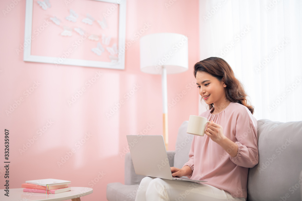 Attractive Asian female lifestyle using the laptop sitting on a couch and drinking coffee Attractive Asian female lifestyle using the laptop sitting on a couch and drinking coffee