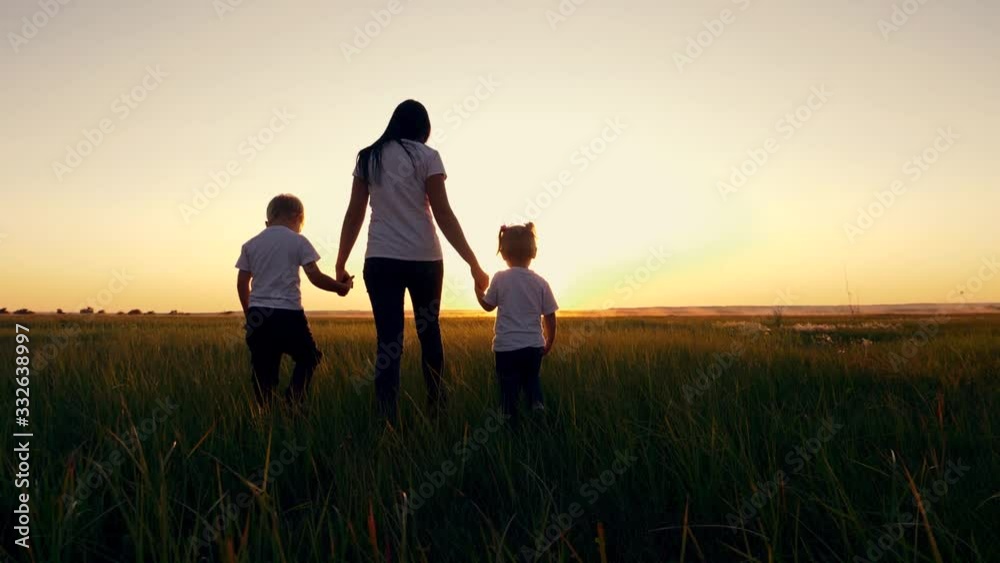 A young woman mother walks with two young children on the grass in the Park. A happy family walks outdoors in the setting sun. A young woman mother walks with two young children on the grass in the Park. A happy family walks outdoors in the setting sun.