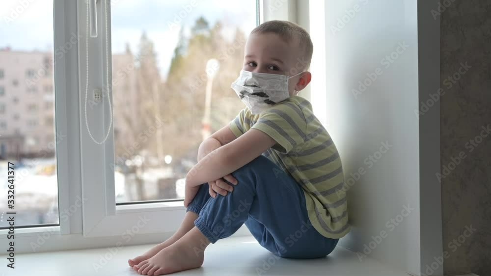 Sad child in protective medical masks lonely sitting near window. Quarantine, staying at home. Slow motion, handheld. Sad child in protective medical masks lonely sitting near window. Quarantine, staying at home. Slow motion, handheld.