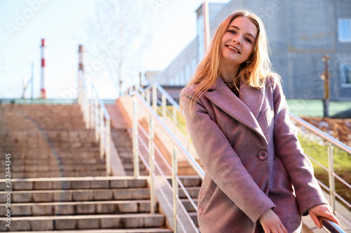 Photo of a cute happy smiling posing blonde Caucasian girl with long beautiful hair outdoors in a pink coat on the background of the steps of the building in sunny day. Model is happy to pose.