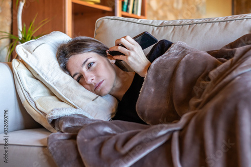 Pretty girl listening to an audio on the smartphone stretched out on the sofa at home covered with a blanket