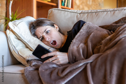 Surprised pretty girl looking at the mobile stretched out on the sofa at home covered with a blanket