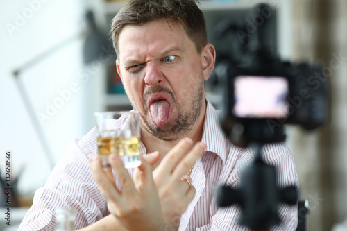 Portrait of man making face holding glass with alcoholic drink. Macro shot of male showing tongue. Videocamera filming on tripod. Beginner blogger concept