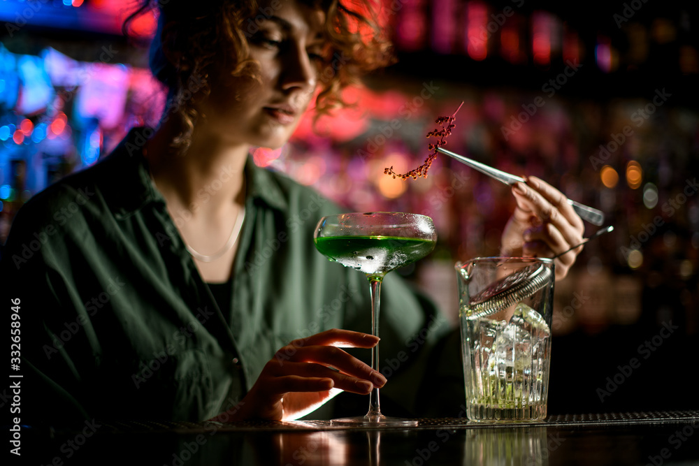 young woman bartender holds glass with cocktail and gently decorates it