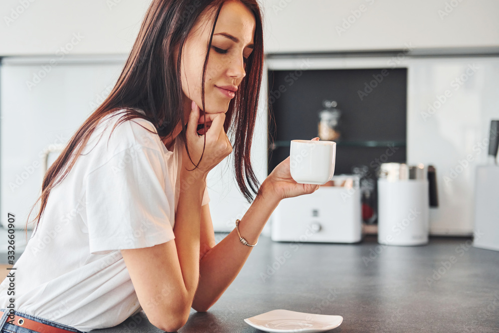 Young beautiful brunette in casual clothes sits in kitchen with fresh drink at morning time Young beautiful brunette in casual clothes sits in kitchen with fresh drink at morning time