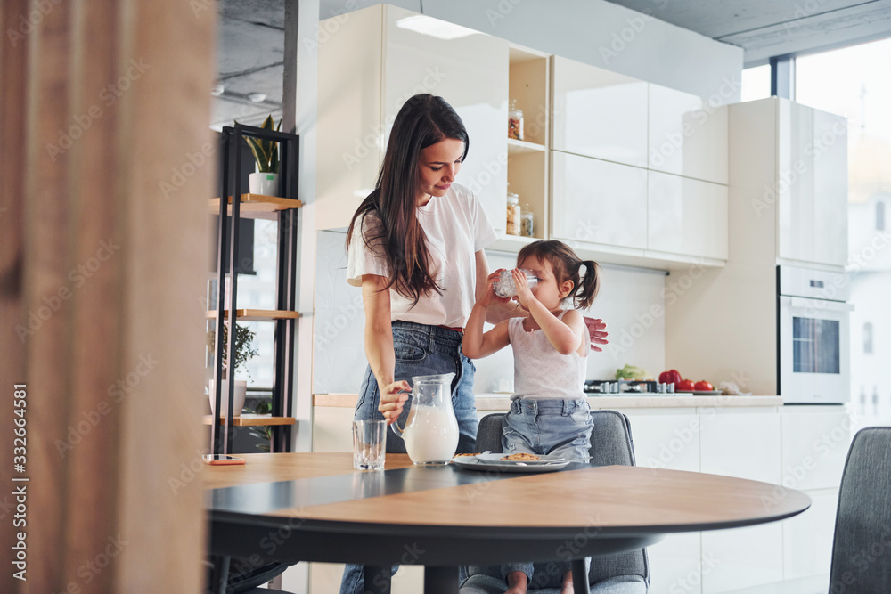 Mother pouring fresh milk into the glass for her little daughter in kitchen Mother pouring fresh milk into the glass for her little daughter in kitchen