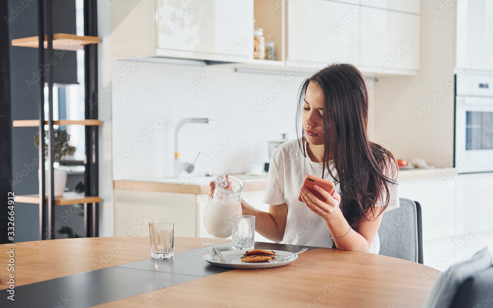 Pouring fresh milk into the glass. Young beautiful brunette in casual clothes indoors in kitchen at daytime Pouring fresh milk into the glass. Young beautiful brunette in casual clothes indoors in kitchen at daytime