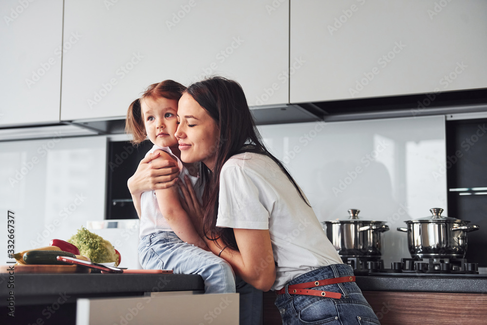 Mother with her little daughter embracing each other indoors in kitchen