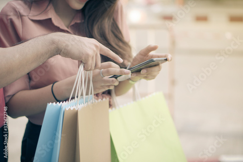 Happy young couple of shoppers walking in the shopping street towards and holding colorful shopping bags in hand and use a smartphone for check promotion. Concept of sale and Black Friday shopping